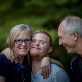 3 people hugging. Female and two males. Adults are grey haired. Teen is blond and appears to have down's syndrome.