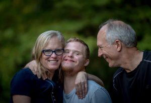 3 people hugging. Female and two males. Adults are grey haired. Teen is blond and appears to have down's syndrome.