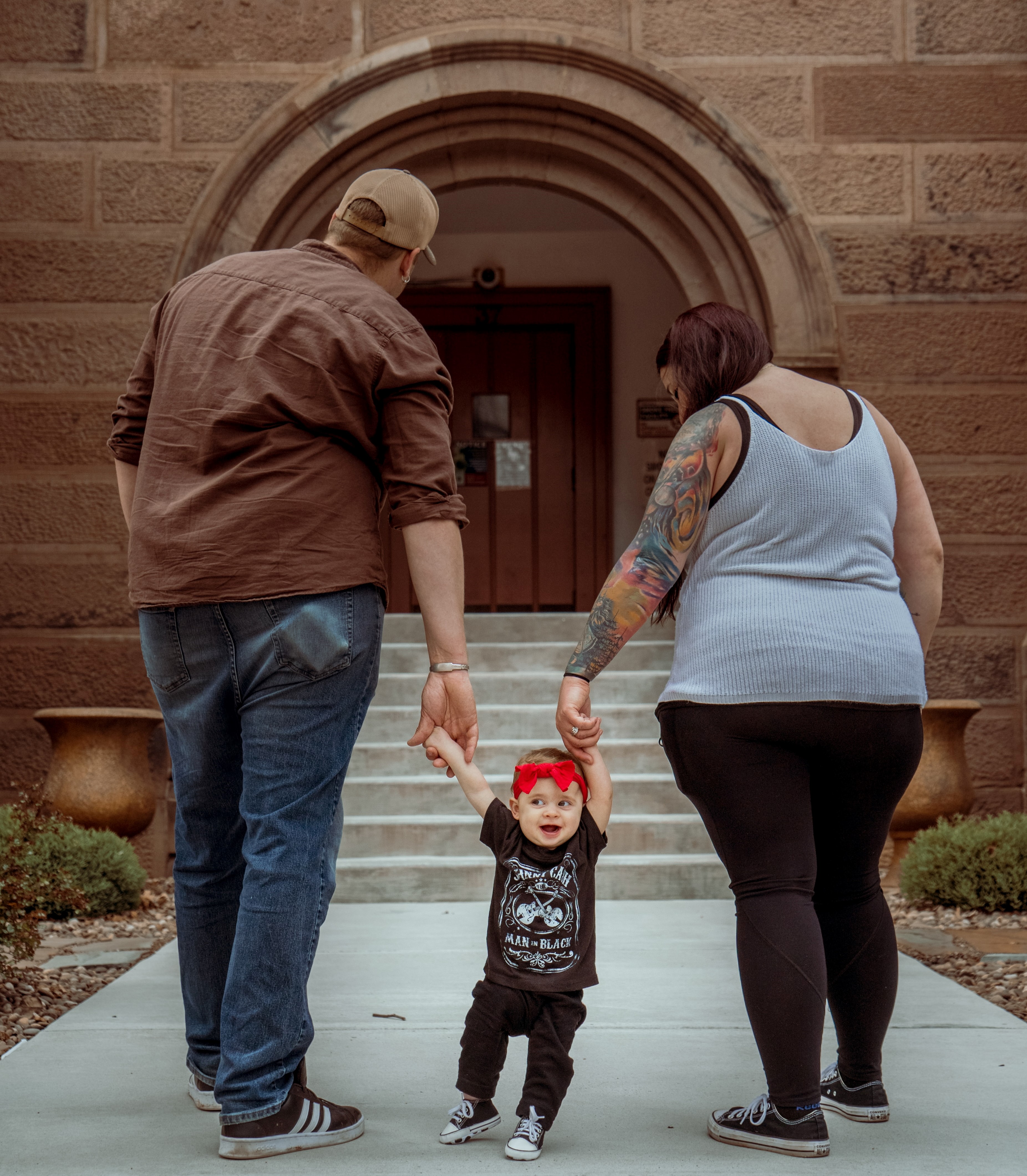 Two parents (appear male and female) holding the hand of a one year old child as they walk toward a government building